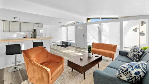 Una sala de estar moderna con un sofá, un sillón y una mesa de centro. Se observa una zona de cocina con taburetes de bar y un ventanal que proporciona luz natural.