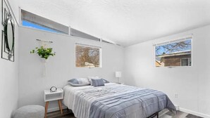 Un dormitorio moderno con una cama, mesitas de noche, una planta colgante y dos ventanas.