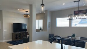 Una cocina moderna con lavavajillas integrado, una zona de comedor con mesa y sillas, televisor e iluminación colgante.