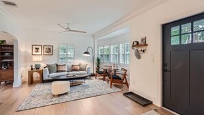 Ein geräumiges Wohnzimmer mit Sofa, Sesseln, einem Couchtisch und einem Teppich. Eine Tür befindet sich rechts, und ein Fenster mit Jalousien ist vorhanden.