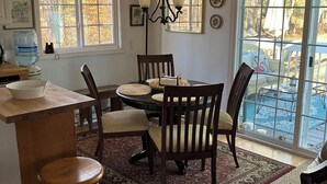 A dining area with a wooden table, chairs, and a chandelier. There are windows with views of trees and a patio area with outdoor furniture.