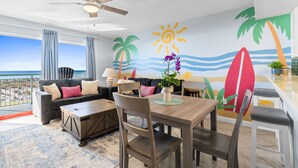 Een kamer met balkon, plafondventilator, eethoek met tafel en stoelen, bank met kussens, en muurschildering met palmbomen en een surfboard.