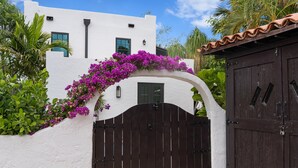 A white building with a dark brown gate, surrounded by lush greenery and purple flowers. - Casa Michaeline Vacation Home (West Palm Beach)