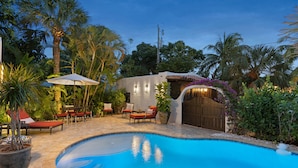 A swimming pool with lounge chairs and umbrellas, surrounded by palm trees and a wooden structure with a decorative arch. - Casa Michaeline Vacation Home (West Palm Beach)