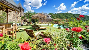 A stone house with a thatched-roof structure, a swimming pool, and lush greenery.
