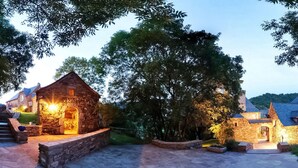 A stone building with a warm light source, surrounded by a stone patio and trees.