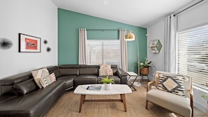 A modern living room with a dark leather sofa, a round coffee table, and a light-colored armchair. There is a ceiling fan, a floor lamp, and a window with blinds.