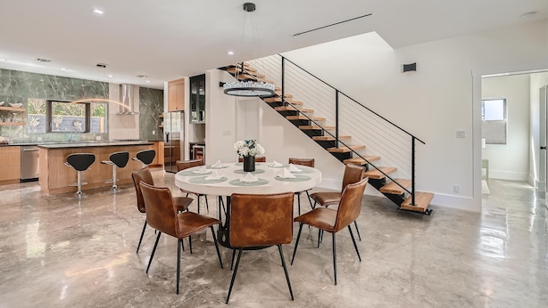 A modern kitchen with a central island, a dining area with a round table and chairs, and a staircase with a wooden finish. - Villa Vogue Vacation Home NEW (West Palm Beach)