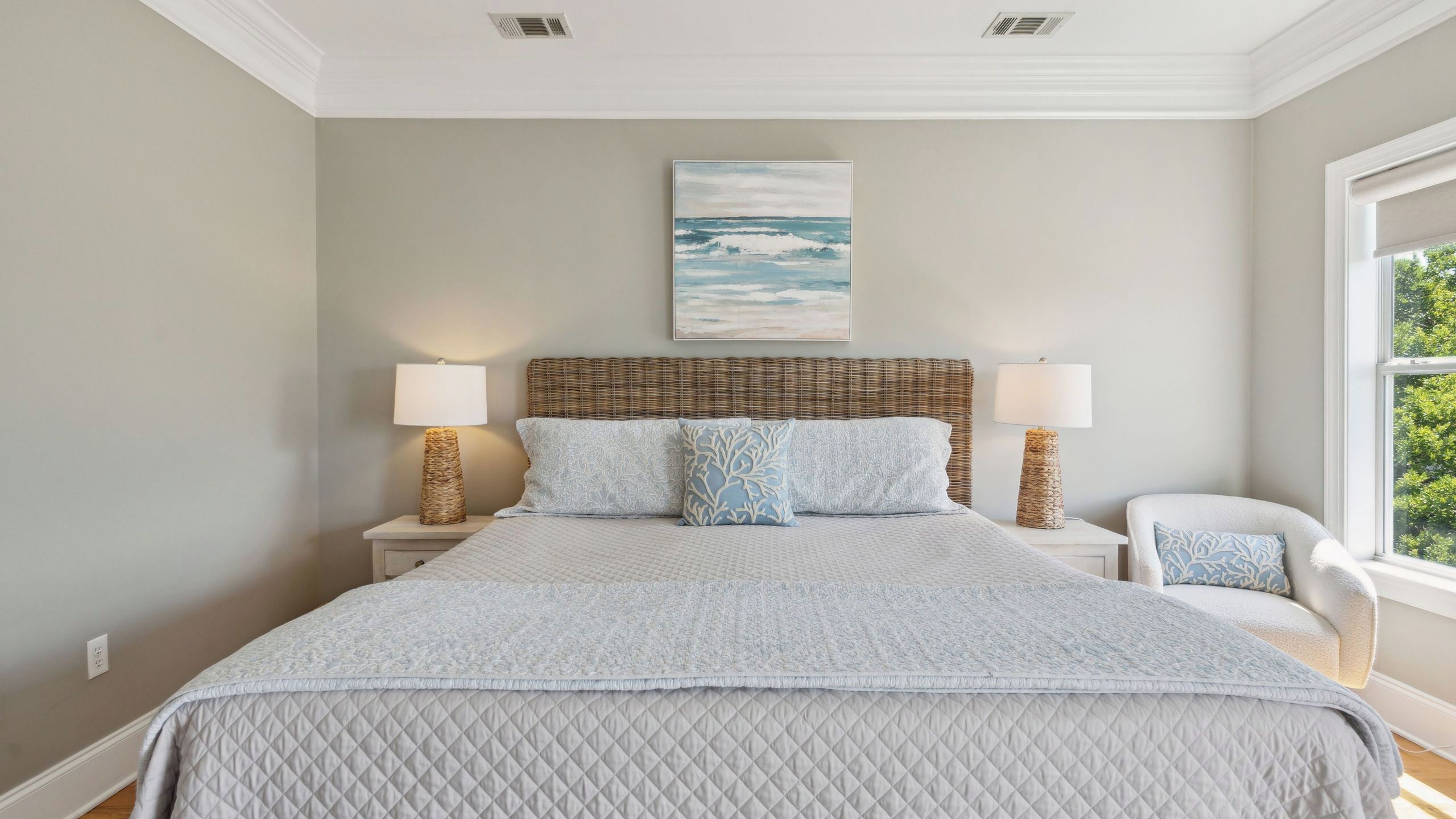 A bedroom with a large bed, two bedside lamps, a wicker headboard, and a painting of the ocean.