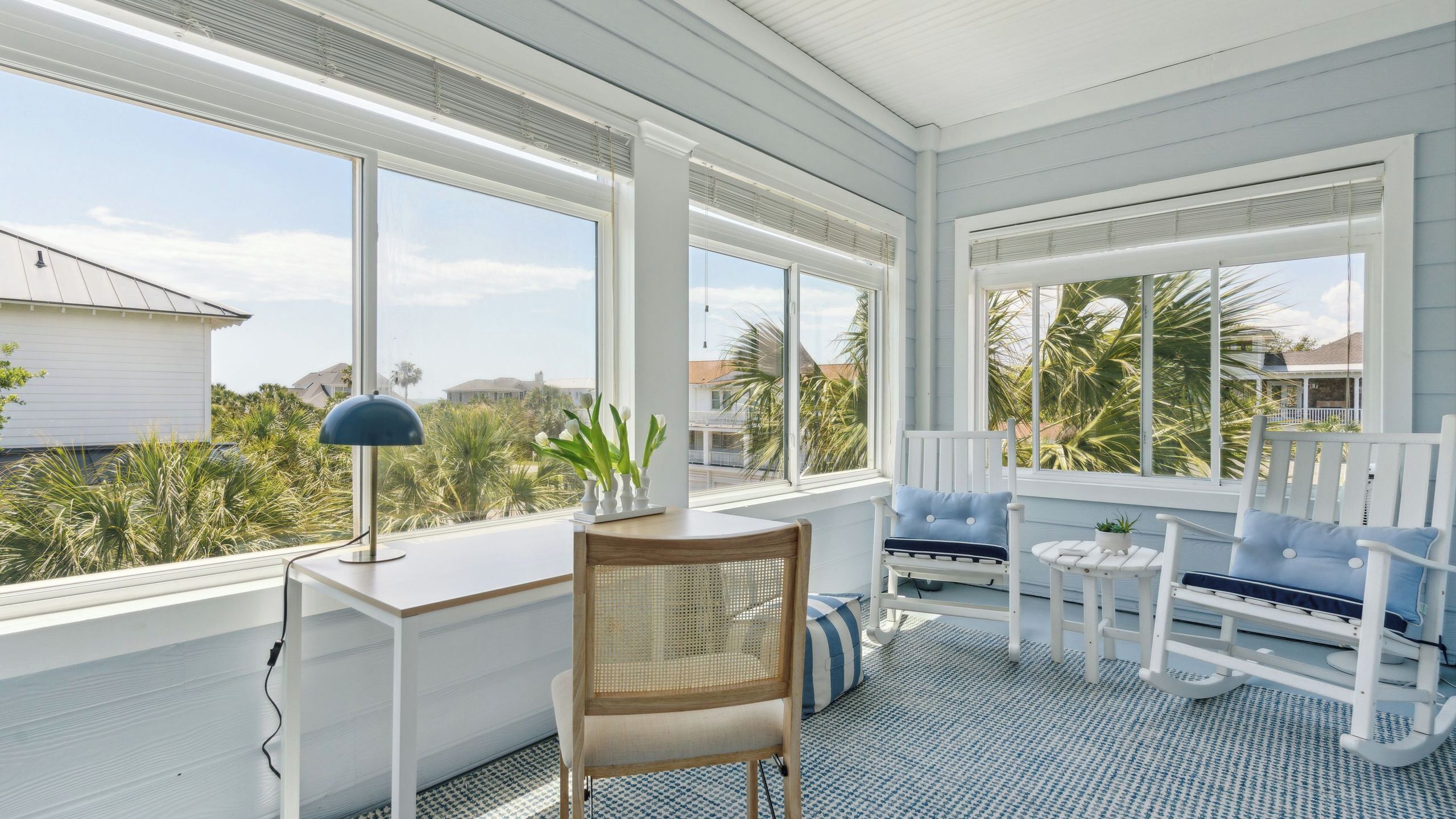 A sunroom with large windows, a desk, a chair, and a seating area with a view of the beach and palm trees.