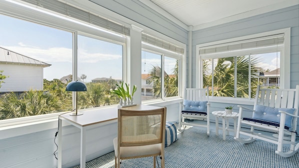 A sunroom with large windows, a desk, a chair, and a seating area with a view of the beach and palm trees.