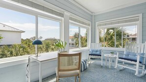 A sunroom with a view of palm trees and houses, a desk with a lamp, and a seating area with cushions. - 3800 Cameron Boulevard Avantstay Pool, Ocean Views, Deck (Isle of Palms)