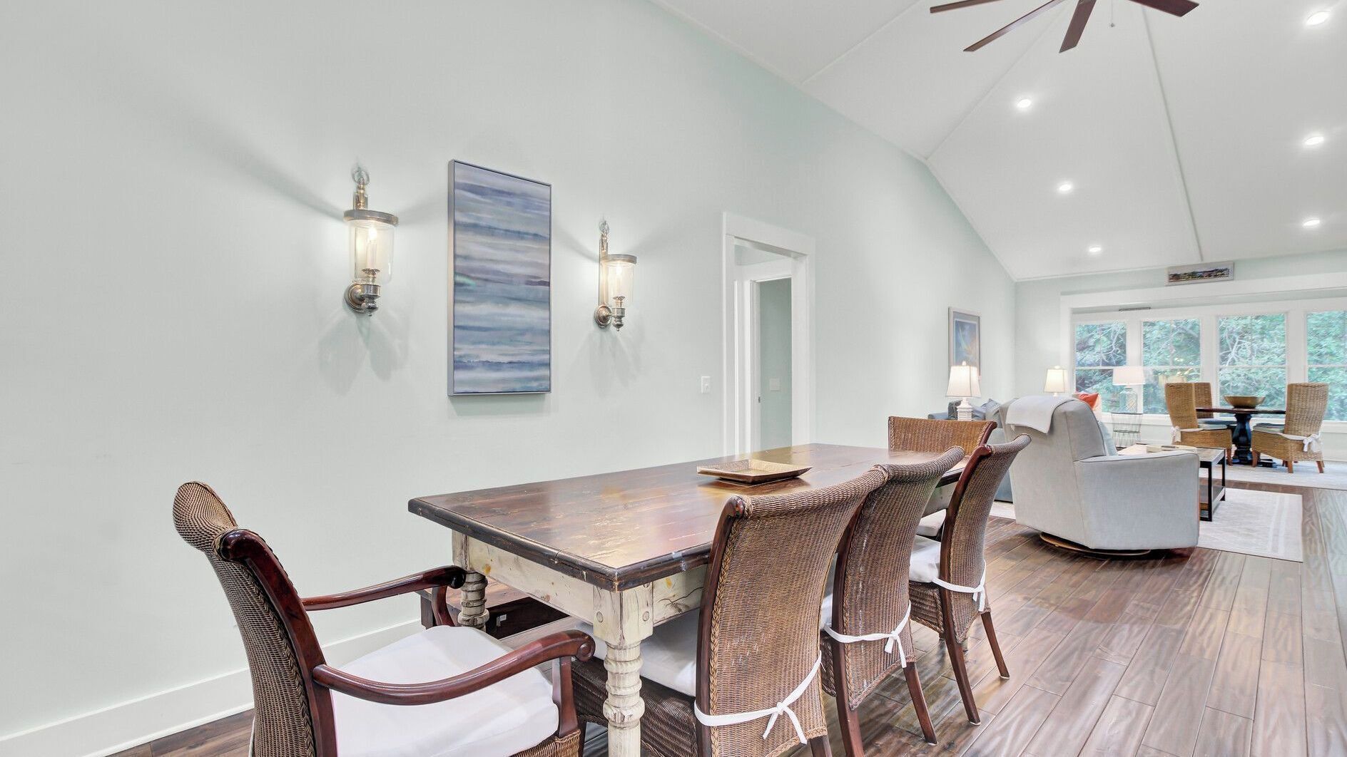 A dining area with a wooden table and wicker chairs, a painting on the wall, and a view of the outdoors through large windows.