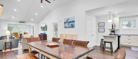 Un comedor amplio con una gran mesa de madera, rodeado de sillas de mimbre, en una habitación luminosa con paredes blancas y un ventilador de techo alto.