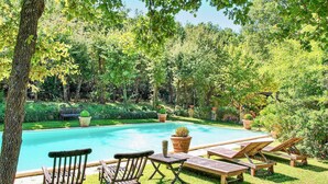 Un área de piscina con sillones reclinables de madera y una pequeña mesa, rodeada de abundante vegetación y árboles.