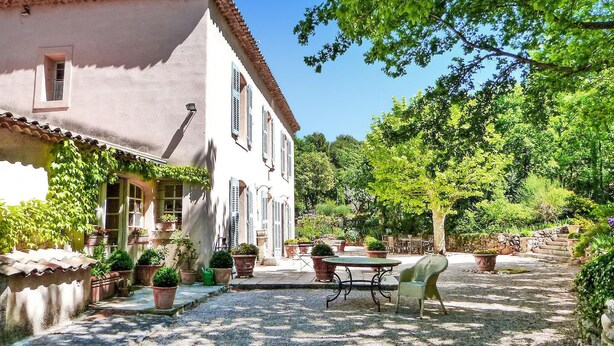 Un patio con una mesa y sillas, rodeado de plantas en macetas y un edificio con contraventanas.
