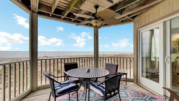 Un espacio al aire libre techado con una mesa y sillas, un ventilador de techo y vista a la playa.