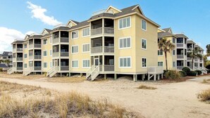 Un hotel frente a la playa con varios pisos, balcones, una playa arenosa y palmeras.
