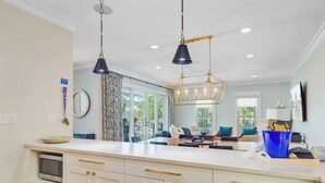 A modern kitchen with white cabinetry, a central island, pendant lighting, and a view of the living area. - 56 Grand Pavilion by Avantstay Oceanfront Deck + Views, Community Pool (Isle of Palms)