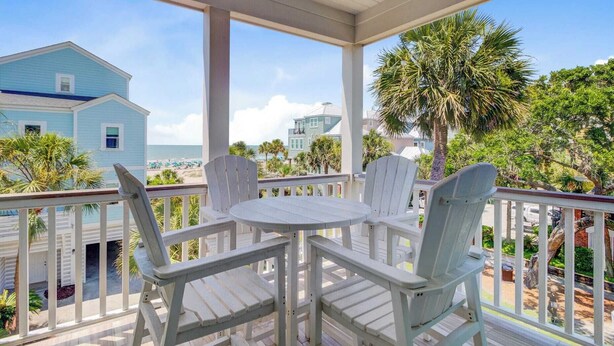 A covered outdoor area with a table and four chairs, overlooking a beach with houses and palm trees. - 56 Grand Pavilion by Avantstay Oceanfront Deck + Views, Community Pool (Isle of Palms)