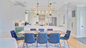 A modern kitchen with white cabinets, a central island, and a dining area with a wooden table and blue chairs. - 56 Grand Pavilion by Avantstay Oceanfront Deck + Views, Community Pool (Isle of Palms)