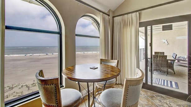 A room with a large window overlooking a beach, a round table with chairs, and a view of the ocean. - 519 Seascape by Avantstay Shared Pool + Views, Incredible Interior Design (Isle of Palms)