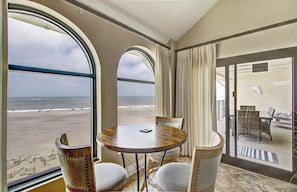 A room with a large window overlooking a beach, a round table with chairs, and a view of the ocean. - 519 Seascape by Avantstay Shared Pool + Views, Incredible Interior Design (Isle of Palms)