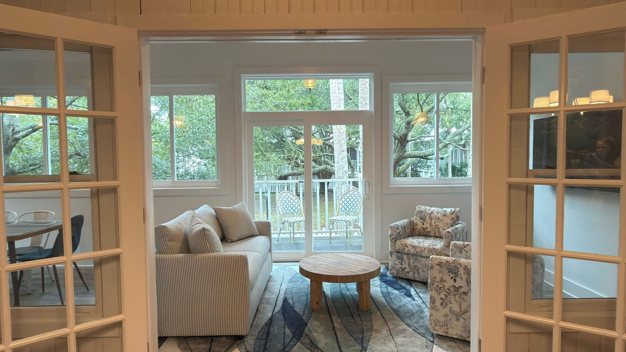 A room with a sofa, two armchairs, a round table, and large windows with a view of trees.