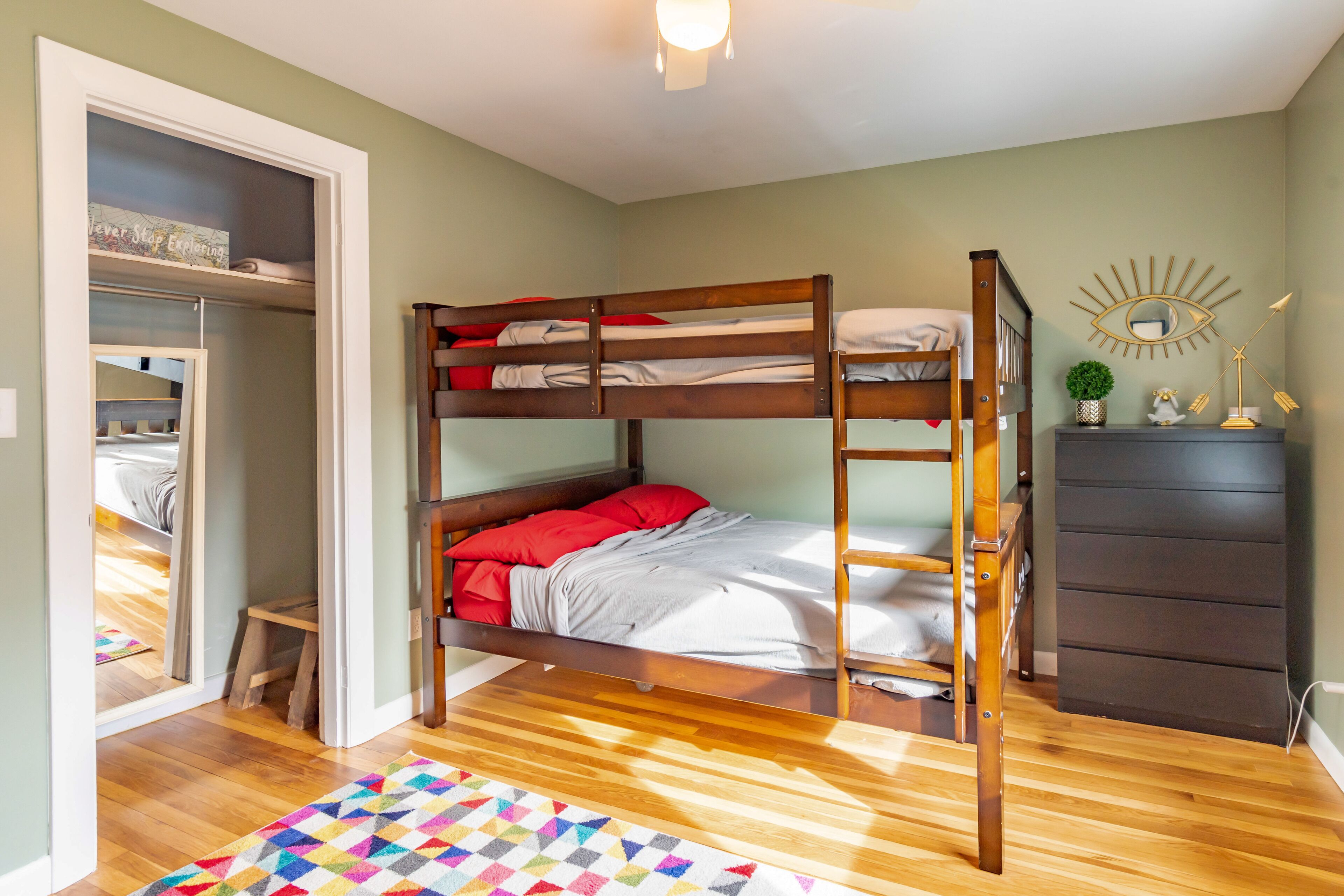 Une chambre avec un lit superposé, un plancher en bois, un petit tabouret, une commode et un miroir au mur.