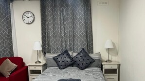 Un dormitori amb un llit blanc, cortines estampades, una cadira vermella i dues taules de nit amb làmpades.