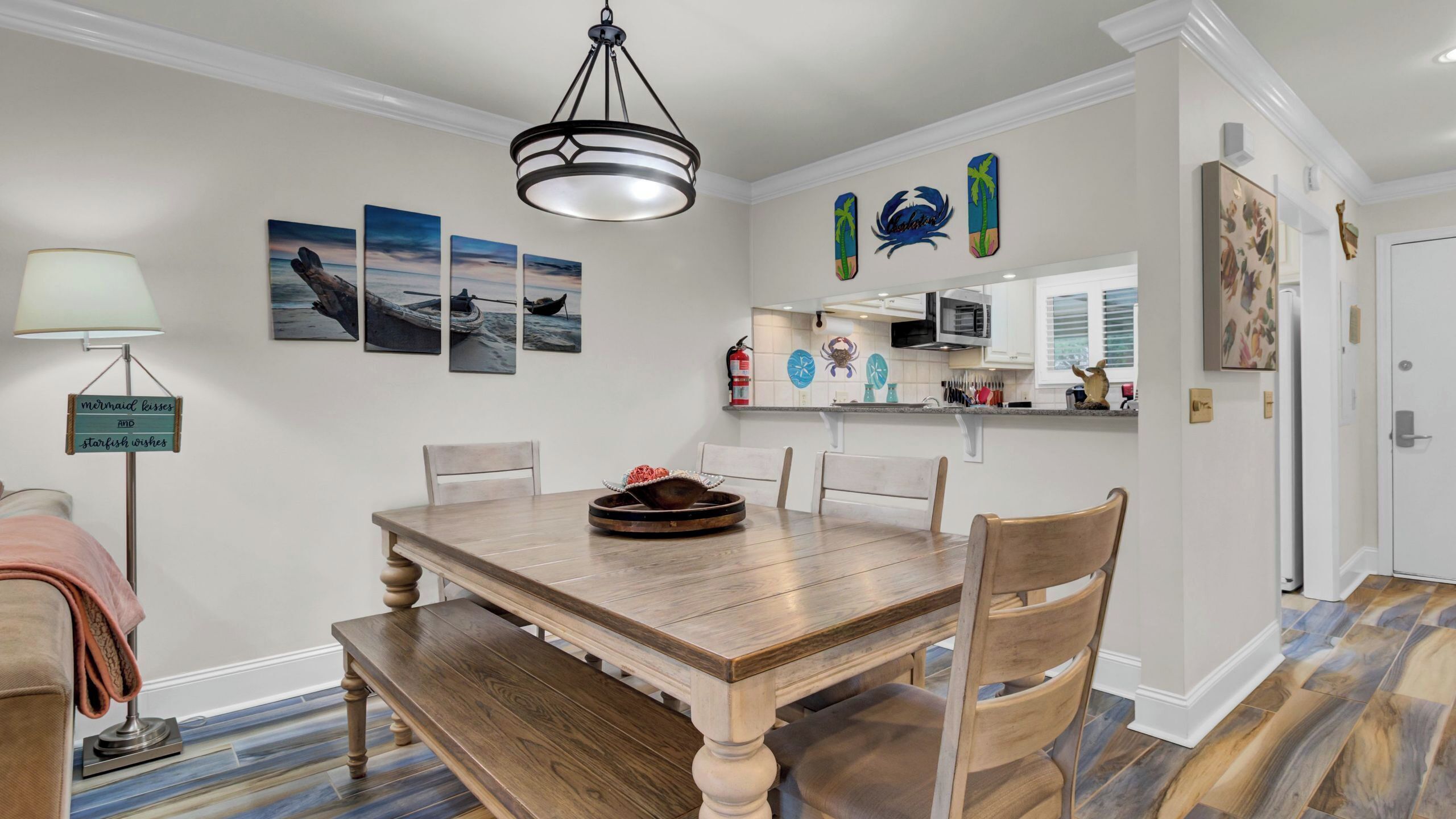 Un comedor con mesa de madera, sillas, un banco, lámpara colgante, decoración en la pared, zona de cocina con armarios y un extintor.