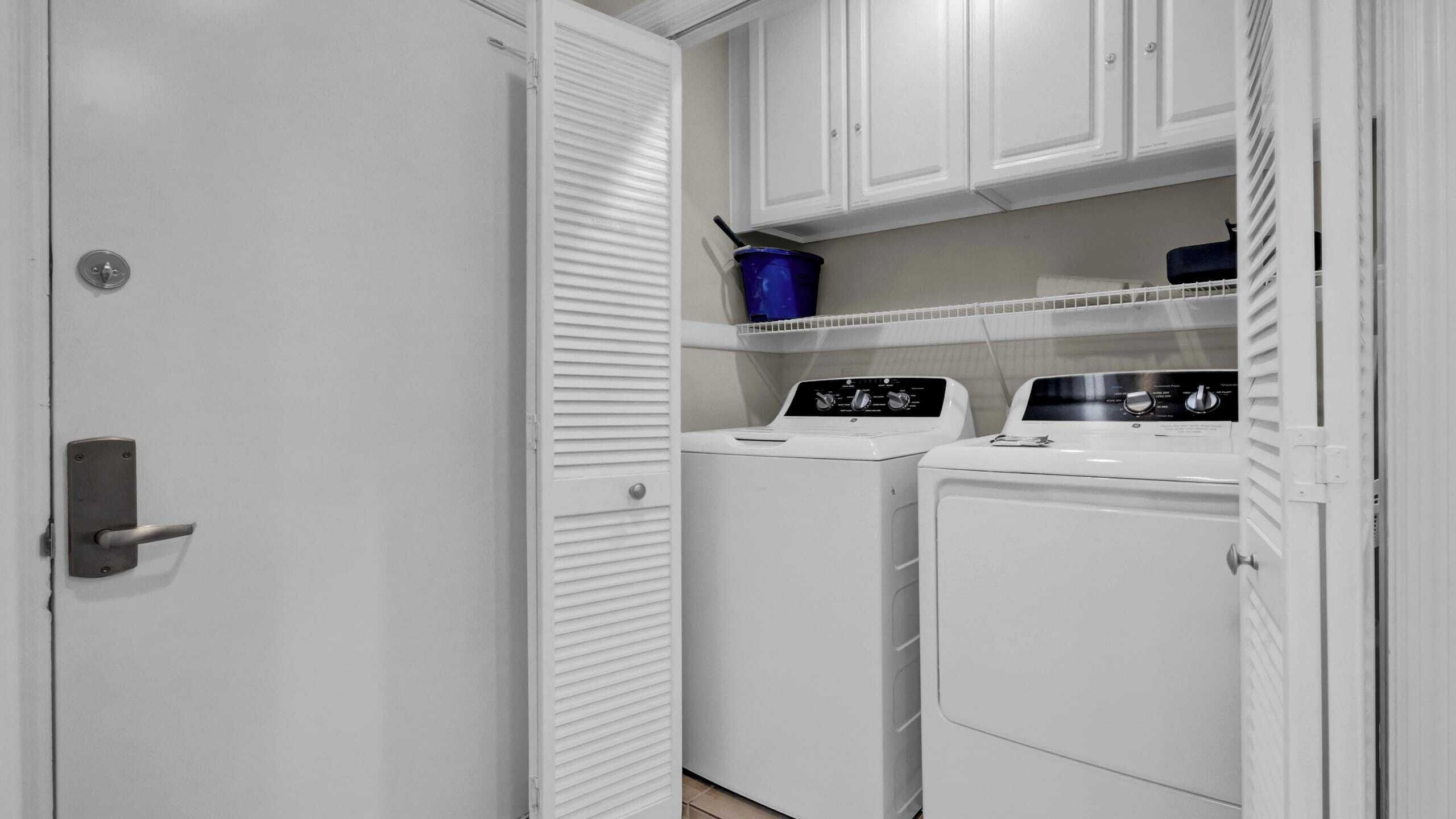 A laundry room with two washing machines and a blue bucket on a shelf.