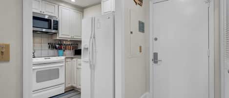A compact kitchen with white cabinets, a microwave, a refrigerator, and a stove. There are knives hanging on the wall and a blue container on the counter.