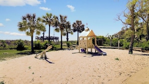 Ein Sandspielplatz mit einer Holzrutsche und Schaukeln, umgeben von Palmen.