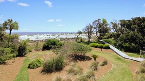 A landscaped garden with a variety of shrubs and trees, a paved walkway, and a view of the beach and ocean in the background. - 121 D Shipwatch by Avantstay Oceanfront Villa w/ View (Isle of Palms)