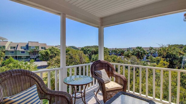 A covered balcony with wicker furniture and a glass table, overlooking a residential area with houses and greenery.