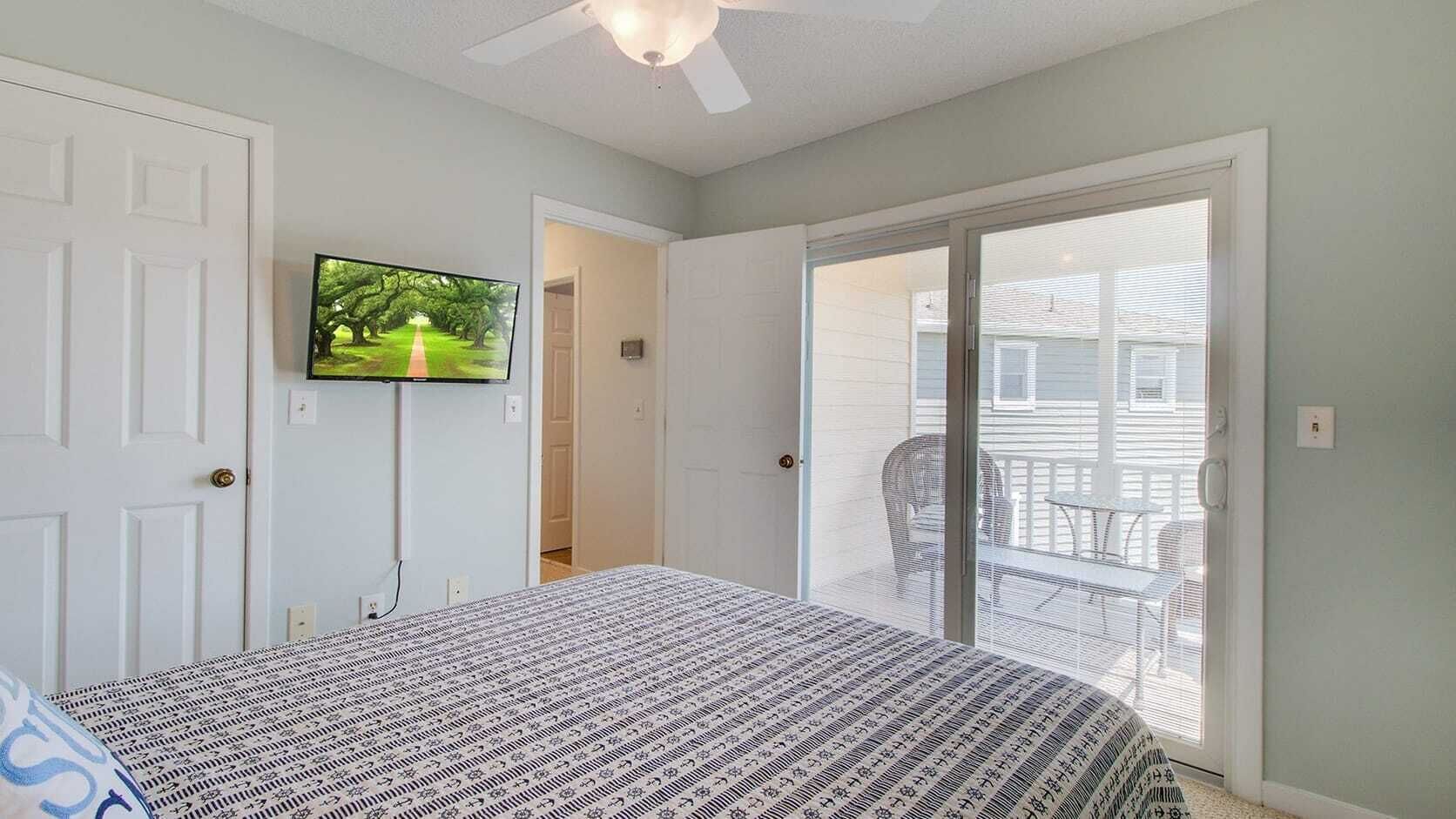 A bedroom with a bed, a television mounted on the wall, and a sliding glass door leading to an outdoor area with a chair and table.