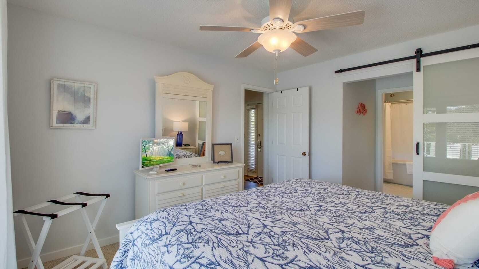 A bedroom with a bed, a dresser, a ceiling fan, a television, and a door leading to another room.