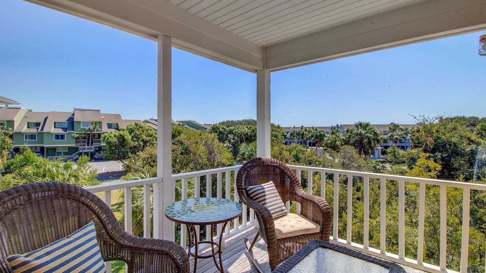 A covered balcony with wicker furniture and a glass table, overlooking a residential area with houses and greenery.