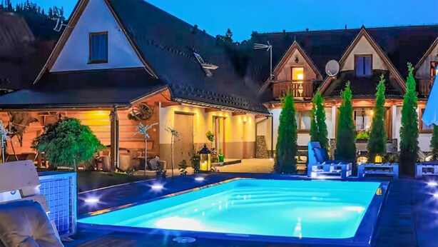 A swimming pool with lounge chairs and a hot tub, surrounded by a wooden deck and a well-lit house with a large window.