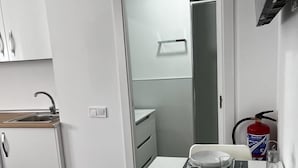 A compact kitchen with white cabinets, a sink, and a dining area with a table set for two. A fire extinguisher is mounted on the wall. - Studio Apartment universidad (Madrid)