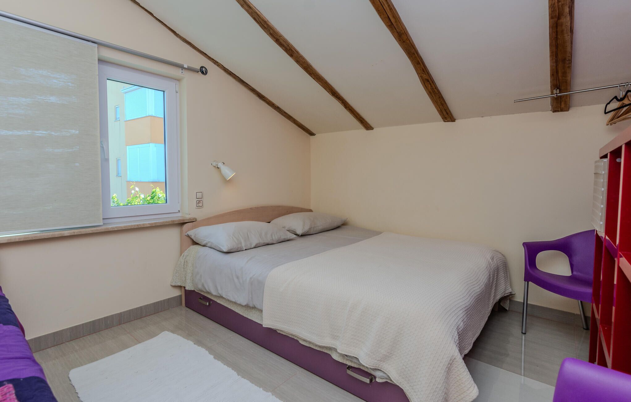 A bedroom with a bed, a window, a wall-mounted lamp, and a chair.