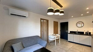 A modern living room with a grey sofa, a small black table, and a white dining table with chairs. There is a kitchen area with a black countertop and white cabinets.