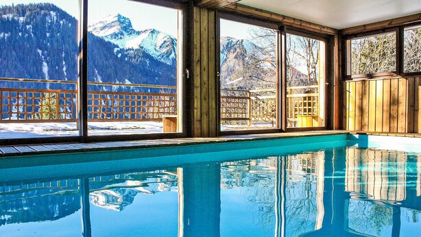 A clear indoor swimming pool with a view of snow-capped mountains through large windows.