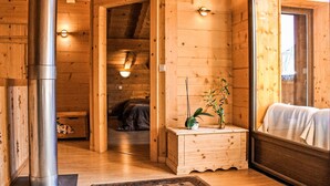 A wooden cabin interior with a central heating pipe, a small wooden cabinet with a plant, and a large mirror reflecting a bed.