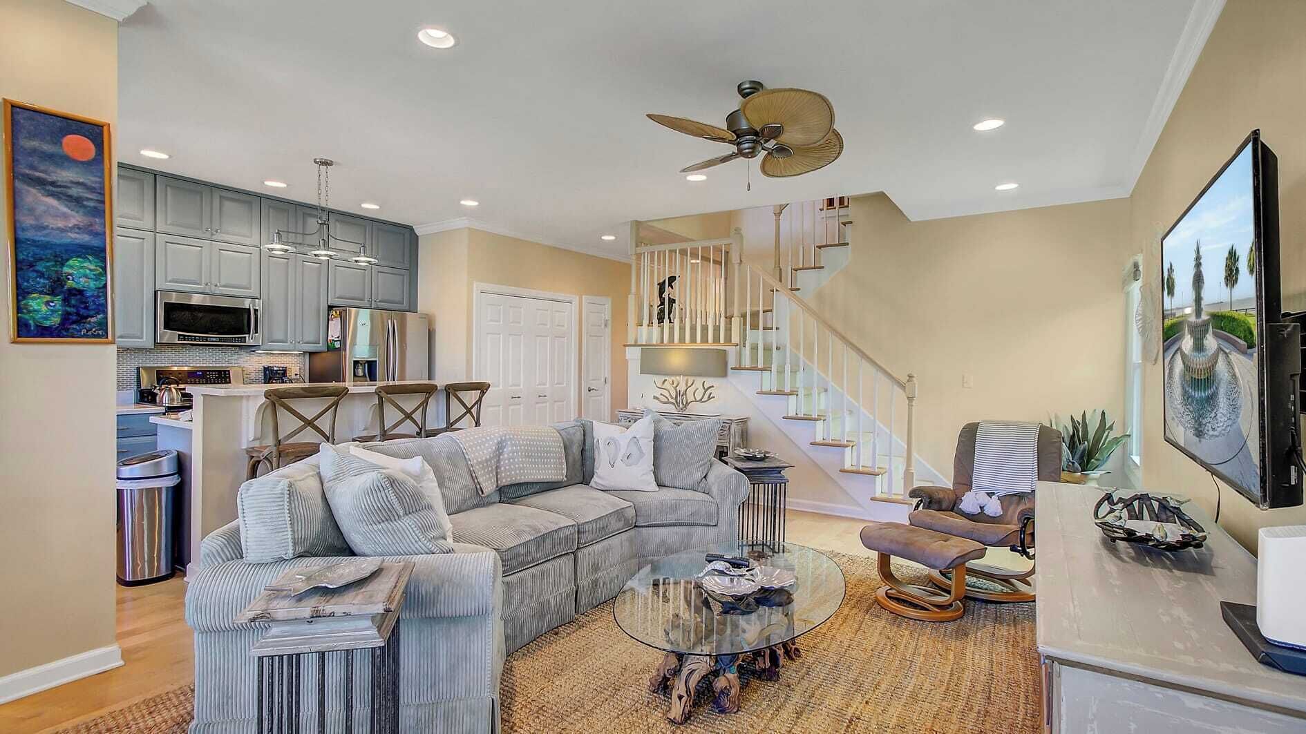 A modern living room with a sectional sofa, a glass coffee table, a fireplace, a staircase, and a kitchen area with cabinets and appliances.