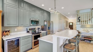 A modern kitchen with grey cabinets, stainless steel appliances, a large island with bar stools, and a chandelier. - 26 Morgans Cove Drive by Avantstay Unique Marina Location (Isle of Palms)