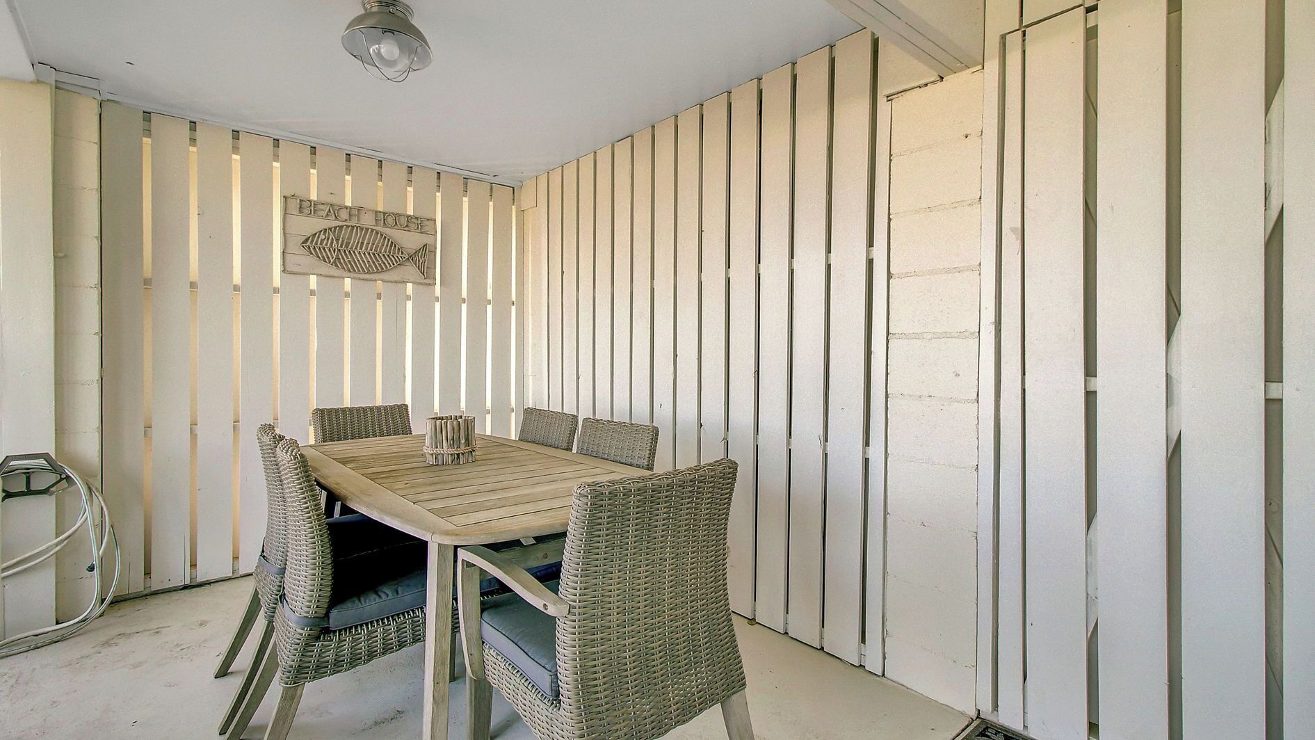 A room with a wooden table and wicker chairs, a ceiling light fixture, and a wall sign reading 'BEACH HOUSE'.