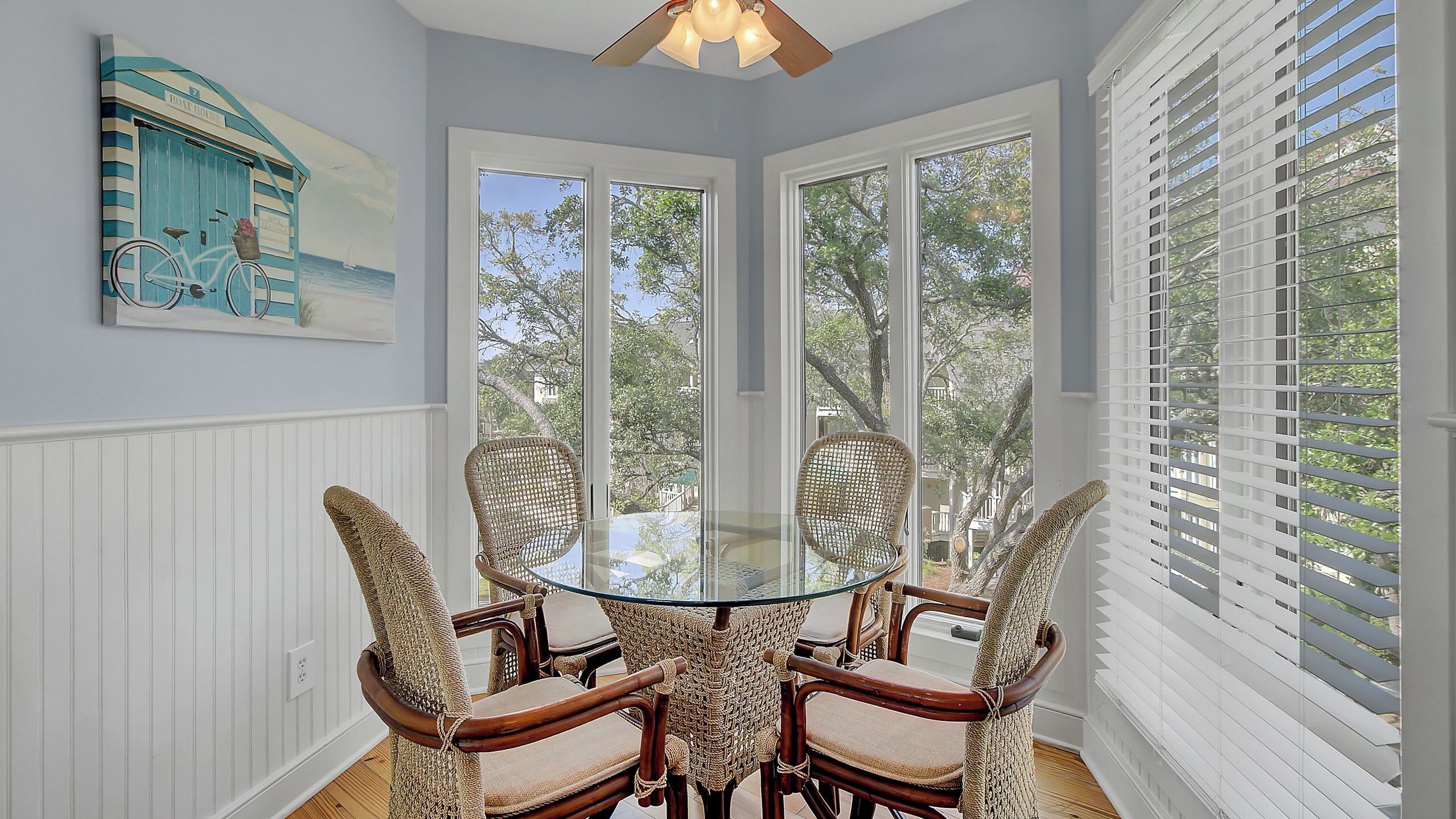 A room with a glass table surrounded by wicker chairs, a ceiling fan, and a view through large windows.