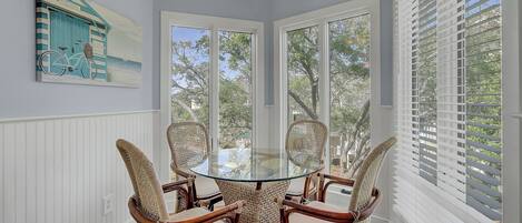 A room with a glass table surrounded by wicker chairs, a ceiling fan, and a view through large windows.